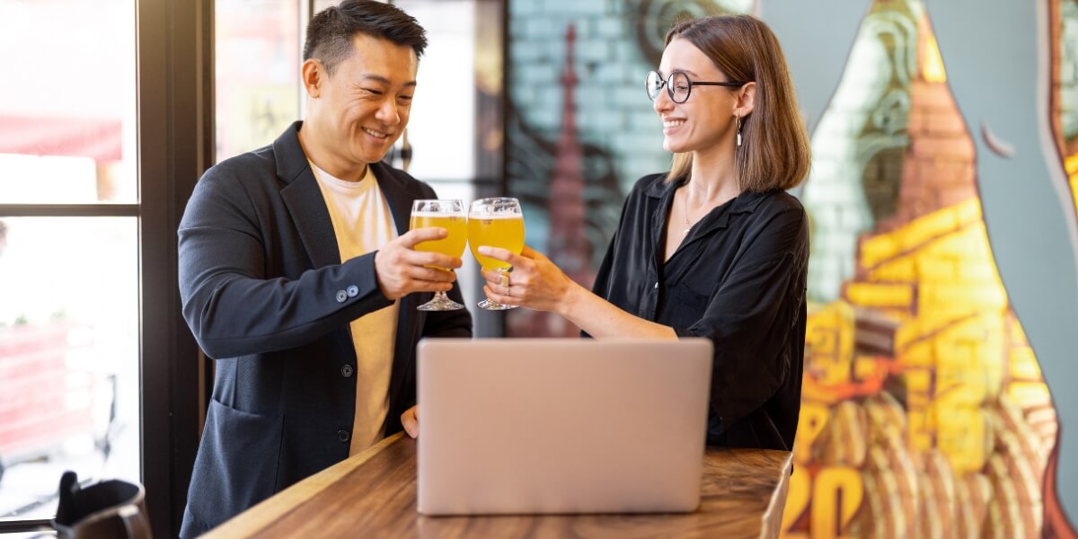 a man and a woman in a bar clinking beer glasses while looking at their brewery business plan on the laptop in front of them