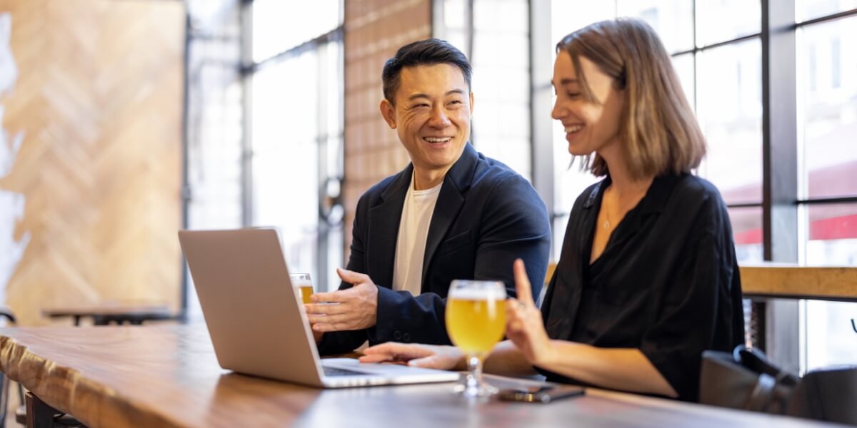 two brewery business partners, a man and a woman at a bar with a laptop sharing a beer while discussing their plans