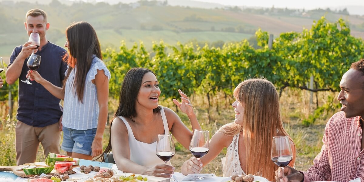 Happy multiracial friends having fun together tasting red wine inside vineyard during summer time