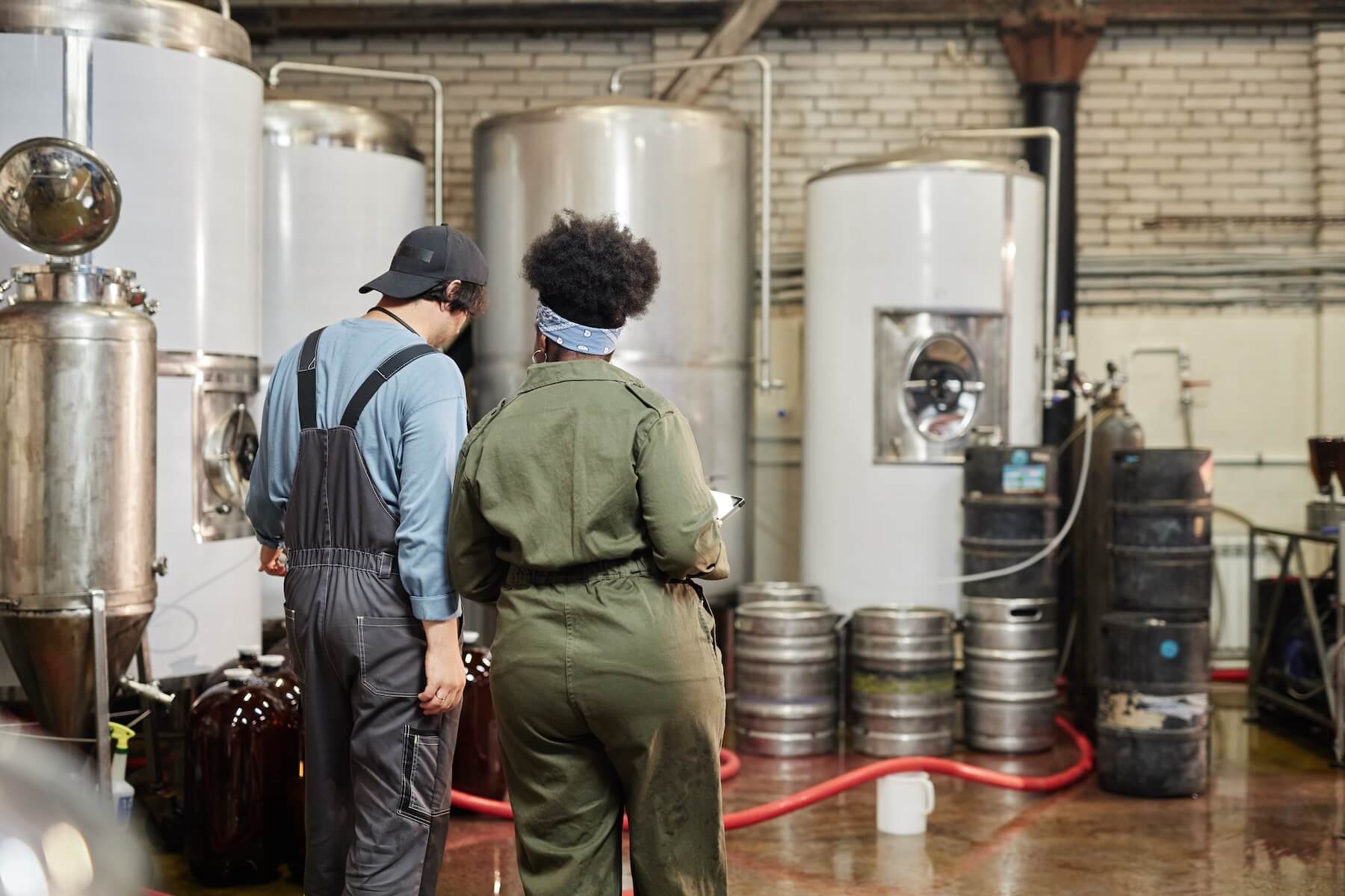 Rear view of diverse team of two brewery workers inspecting production process with huge fermentation tanks