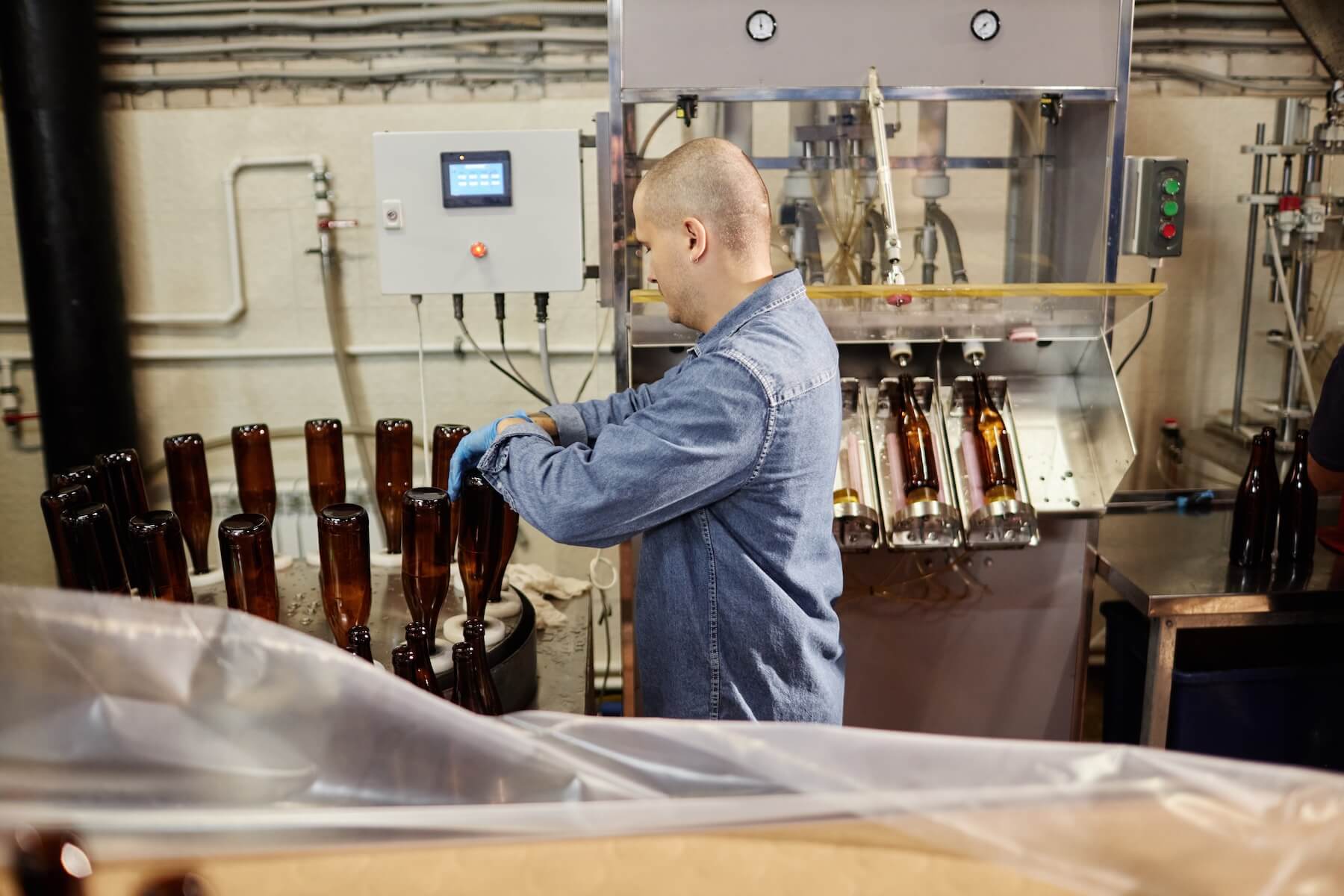 Side view of concentrated male production operator carefully sterilizing glass bottles while directing cleaning machine in workshop of cider or beer production facility