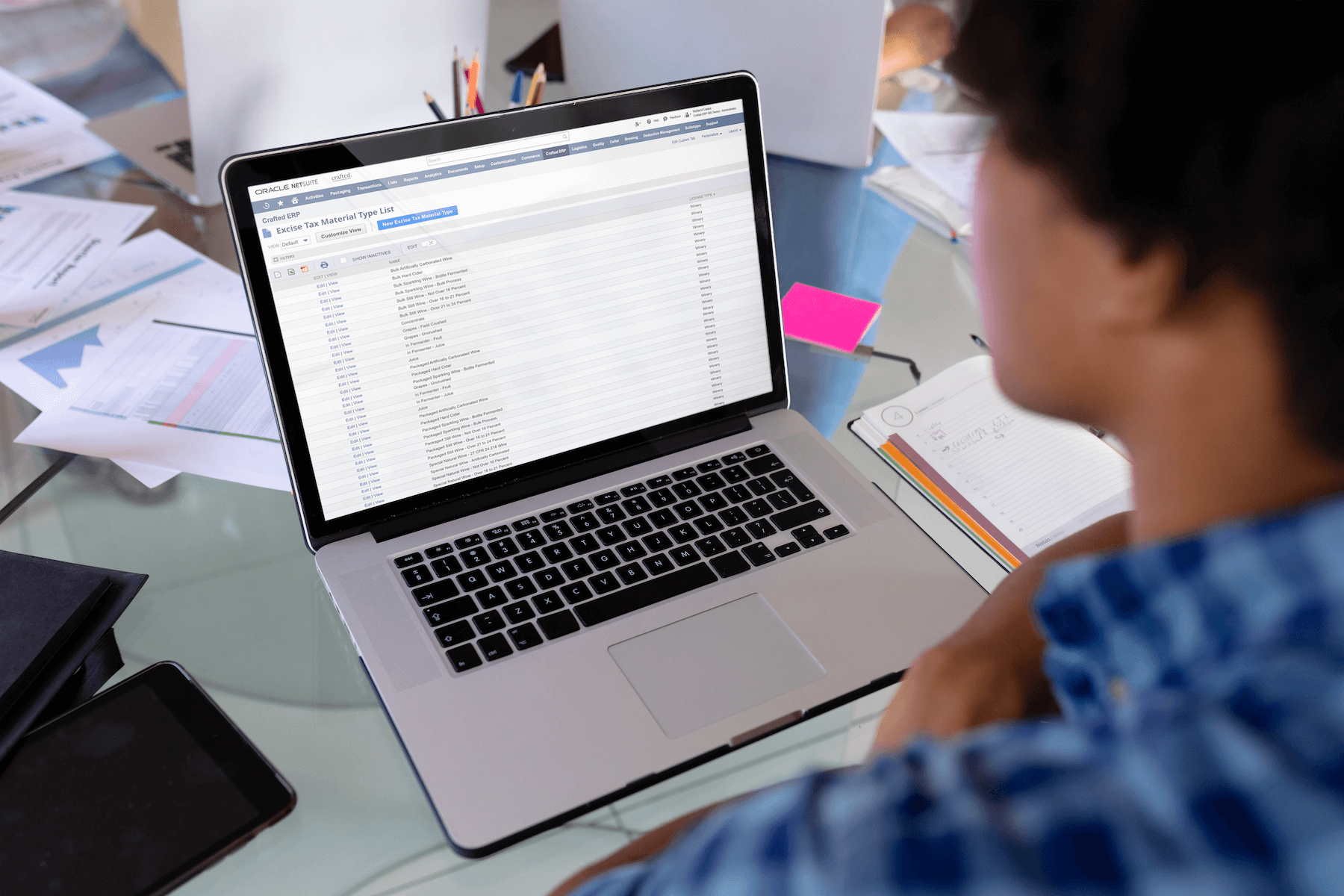An African American woman working on a laptop at a winery. She is managing winery compliance and looking at an excise tax materials list.