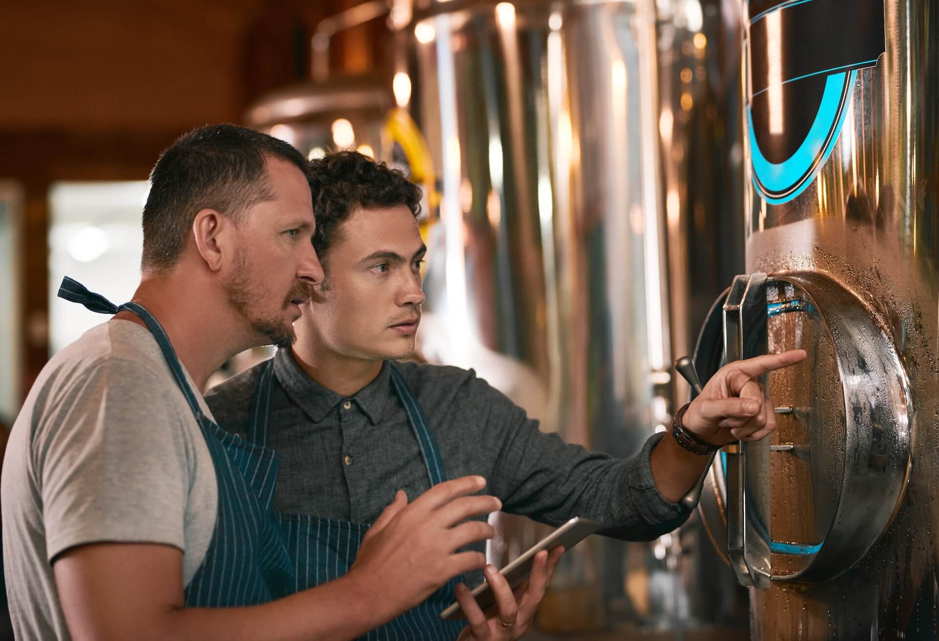 Two brewers looking at tanks and entering data on a tablet in a beverage ERP software platform